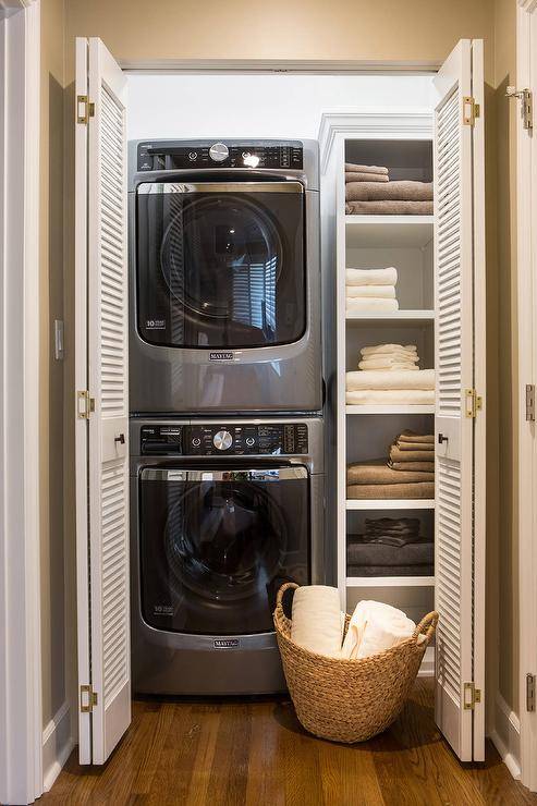 A Maytag washer and dryer is stacked in this organized closet with folding louvered doors. This humble vertical space features built in towel and linen shelves that creates a practical space saving laundry room.