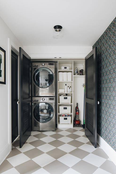 Kate Marker Interiors - Stoffer Photography - A stacked silver washer and dryer sits on white and gray diamond pattern floor tiles behind black shutter doors and next to white shelves lit by a clear glass flush mount.