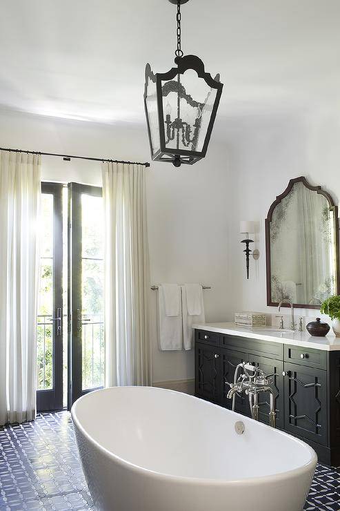 Black French patio doors covered with ivory curtains hung from an oil rubbed bronze curtain rug allow natural light to stream in to a black and white Mediterranean style bathroom fitted with a freestanding oval bathtub placed in the center of the room on black and white mosaic floor tiles beneath an iron and seeded glass lantern. The tub is paired with a polished nickel floor mount tub filler fixed in front of a black washstand complemented with black star cabinet doors contrasted with white knobs and an ivory quartz countertop paired with an ivory quartz backsplash fixed behind a satin nickel gooseneck faucet. A brown arch mirror is mounted over the washstand between iron single light sconces.