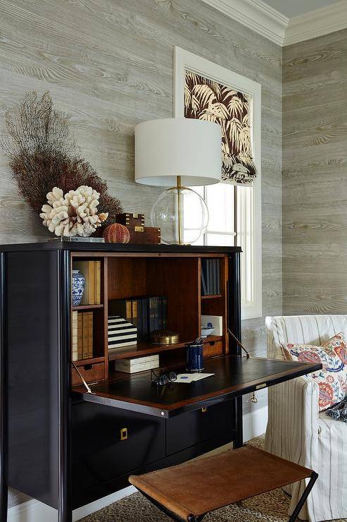 A brown leather stool is paired with a black secretary desk adorned with square brass drop pulls topped with sea coral, sea fan and an Aerin Morton Table Lamp lining a wall clad in gray faux bois wallpaper.