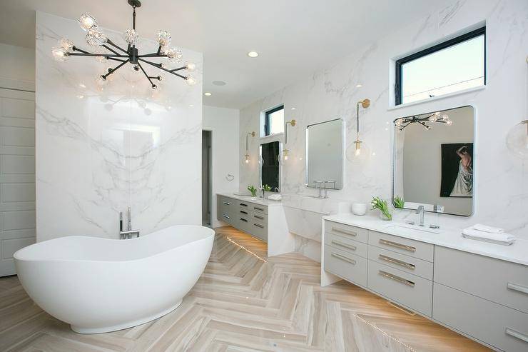master bathroom boasting a modern freestanding bathtub placed on herringbone floor tiles beneath a black modular chandelier hung in front of a marble clad wall. His and her light gray washstands are custom lit and adorn long polished nickel pulls and white quartz countertops. The countertops are fitted with sinks and modern polished nickel faucet kits fixed beneath Restoration Hardware Bristol Flat Mirrors mounted below clerestory windows and illumi