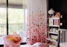 Pink paint splatter curtains cover glass sliding doors and pair well with a black accent wall fixed behind a two tone bookcase placed on a cream diamond rug beside a black crib. Pink velvet pillows are placed on a white sheepskin rug positioned in front of a pink accent chair.