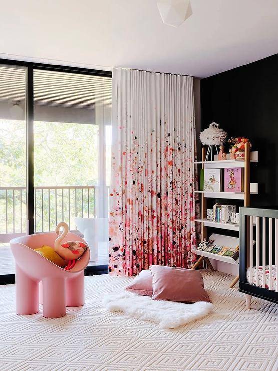 Pink paint splatter curtains cover glass sliding doors and pair well with a black accent wall fixed behind a two tone bookcase placed on a cream diamond rug beside a black crib. Pink velvet pillows are placed on a white sheepskin rug positioned in front of a pink accent chair.