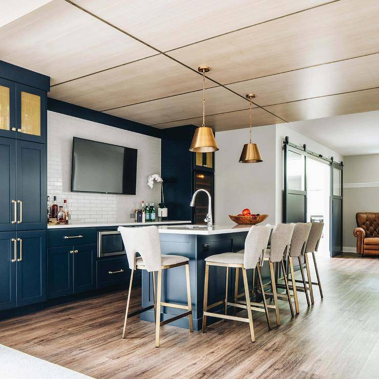 Two brass drum pendants hang over a blue kitchenette island matched with light gray linen counter stools placed at a white quartz countertop holding a sink with a polished nickel gooseneck faucet. A flat panel TV is mounted to white brick tiles between peacock blue and gold cabinets stacked over peacock bleu cabinets and a glass front wine fridge.