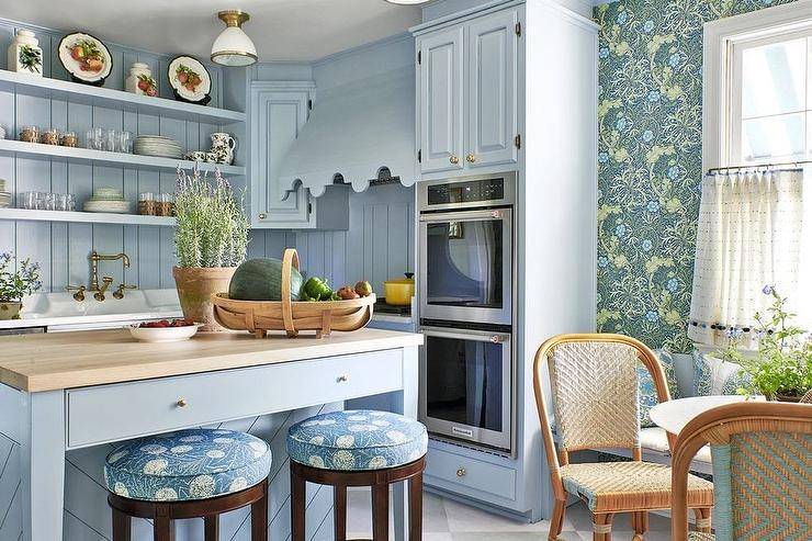 Lovely blue cottage kitchen features a blue chevron island topped with a butcher block countertop and paired with round brown wood stools with blue cushions. Blue shelves are stacked against a blue plank backsplash over a sink with a brass hook and spout faucet, while a dual wall oven is positioned under blue cabinets and beside a blue scalloped range hood.