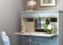 French blue secretaire desk positioned against a gray wall and beneath a gold framed art piece complete with matching chair