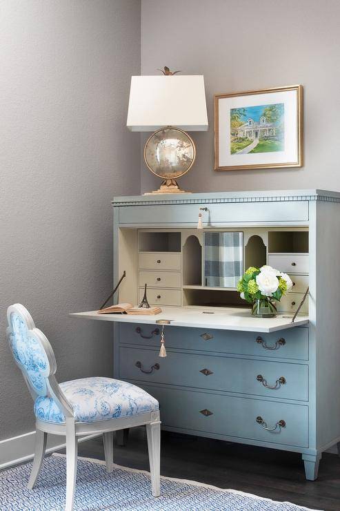 French blue secretaire desk positioned against a gray wall and beneath a gold framed art piece complete with matching chair