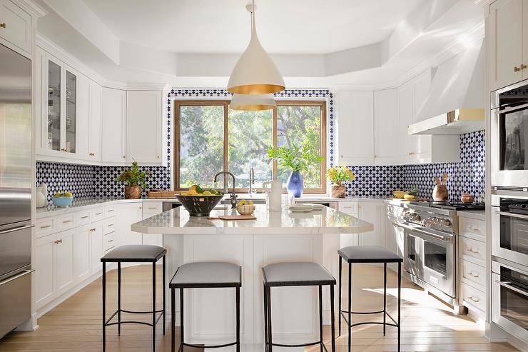 White and gold lanterns hung from a tray ceiling illuminate a white shaker island topped with a marble countertop finished with a prep sink and paired with backless gray and black counter stools. White perimeter cabinets are complemented with white and blue mosaic backsplash tiles.