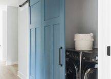 White hallway with blue barn doors on rails opening into a laundry closet featuring a silver front load washer and dryer. A black iron geometric lantern illuminates the hallway above blond hardwood floors.