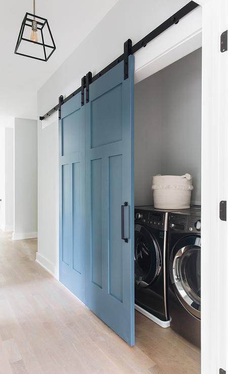 White hallway with blue barn doors on rails opening into a laundry closet featuring a silver front load washer and dryer. A black iron geometric lantern illuminates the hallway above blond hardwood floors.