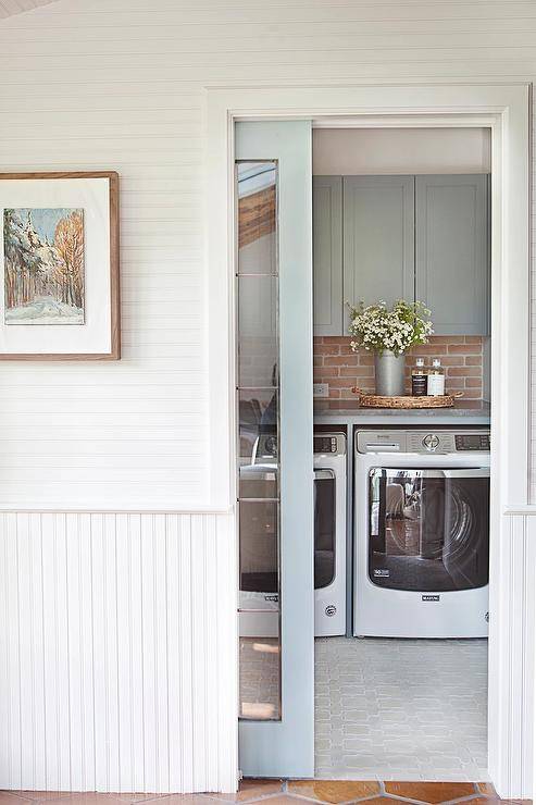 Blue sliding door with glass panels opens to a blue laundry room featuring an enclosed white front loading washer and dryer placed on light gray floor tiles under blue cabinets mounted against a red brick backsplash.