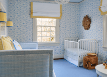 Sweet yellow and blue nursery is clad in blue wallpaper and boasts windows covered in yellow grosgrain roman shades hung under a blue ceiling. A woven elephant stool sits on a blue rug beside a white vintage metal crib, while yellow and blue sconces flank an art piece hung above a blue daybed topped with yellow pillows.