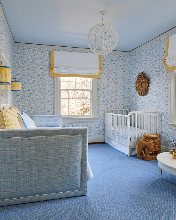 Sweet yellow and blue nursery is clad in blue wallpaper and boasts windows covered in yellow grosgrain roman shades hung under a blue ceiling. A woven elephant stool sits on a blue rug beside a white vintage metal crib, while yellow and blue sconces flank an art piece hung above a blue daybed topped with yellow pillows.