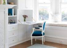 Beside a white built-in bay window bench topped with a natural linen cushion, white built in cabinets donning brass knobs feature a drop desk located in front of a blue velvet French Louis chair.
