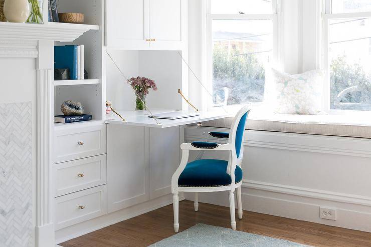 Beside a white built-in bay window bench topped with a natural linen cushion, white built in cabinets donning brass knobs feature a drop desk located in front of a blue velvet French Louis chair.