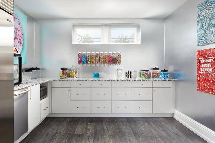 Contemporary candy themed kitchenette in basement features wall mount candy dispensers and candy art on silver foil wallpaper, candy jars on black and white marble countertop over white cabinets with glass and brass pulls and dark wooden floors.