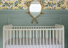 A striking faux bois mirror hangs over a vintage white spindle crib in front of a green wainscot wall finished with lemon print wallpaper and green crown moldings.