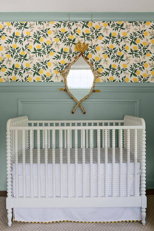 A striking faux bois mirror hangs over a vintage white spindle crib in front of a green wainscot wall finished with lemon print wallpaper and green crown moldings.