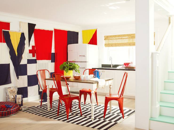 Burnham Design - Fabulous basement features a wall covered in flags beside kitchenette filled with Big Chill Refrigerator beside white cabinets topped with black countertop framing sink under window dressed in bamboo roller shades. A farmhouse dining table is lined with red metal dining chairs atop a World Market Black and White Striped Dhurrie Rug.