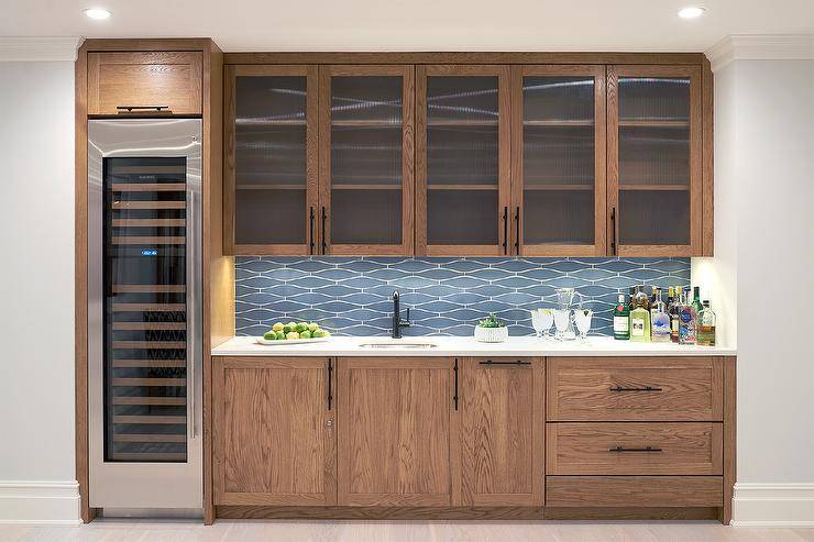 This gorgeous basement boasts a wet bar fitted with brown oak cabinets topped with a white quartz countertop finished with a sink and an oil rubbed bronze gooseneck faucet. The faucet is mounted in front of blue geometric backsplash tiles and beneath glass front cabinets. A tall wine fridge is recessed beneath a single brown oak cabinet.