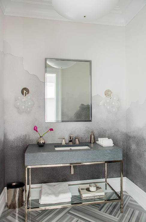 A nickel framed mirror hangs from a wall covered in a white and gray mural and between white glass globe sconces lighting gray marble herringbone floor tiles. Beneath the mirror, a stone and nickel sink vanity is fitted with a glass shelf and a polished nickel faucet.