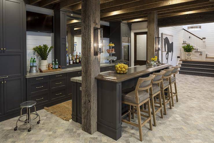 Cabin style basement with a wet bar showcasing black fabric and wood barstools at a dark gray island with concrete countertops. Shiplap backsplash lifts the lighter neutrals while warm hues fill the wet bar with dark cabinets, stone floors, and rustic wood support beams.