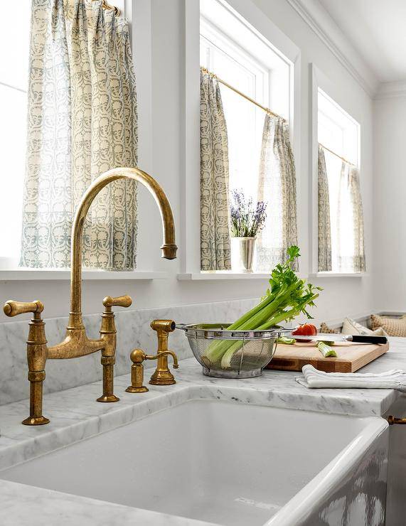 Cottage style kitchen features an aged brass deck mount faucet with gray marble countertop, a farm sink and yellow and gray cafe curtains.