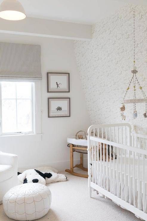 Lovely monochromatic nursery boasts a sheep mobile hung in front of an vocalizing wall covered in white and gray horse print wallpaper and over a white crib. The crib is accented with a white ruffled skirt and placed on ivory carpeting. Stacked art hangs abreast a window covered in a stripe gray roman shade.