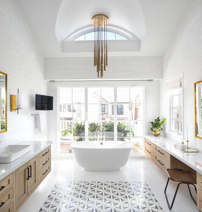 master bathroom is lit by gold staggered lights hung over an oval freestanding bathtub paired with a polished nickel tub filler mounted in front of windows, while a flat panel television is fixed in a corner over a polished nickel towel bar. White floor tiles surround white and gray geometric accent tiles flanked by a long gray wash makeup vanity fitted with a white marble countertop mounted under an RH Modern Pendant Mirror and a gray wash wood washstand. The washstand is finished with oil rubbed bronze pulls and a white marble countertop holding a rectangular vessel sink under a RH Modern Pendant Mirror lit by a brass and white glass sconce mounted to white subway wall tiles.