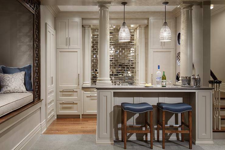 Chic basement bar features Aesthetic Muse pendants illuminating a built-in bar topped with grey quartz and lined with backless blue leather barstools with silver nailhead trim. Basement bar boasts ivory shaker cabinets paired with gray quartz counters and an beveled mirrored subway tiled backsplash from The Fine Line Tiles flanked by paneled refrigerators. Basement is fitted with a built-in reading nook filled with gray and blue cushions finished with a wood lattice frame.