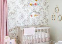 A brass lantern illuminates a stunning pink, white, and gray nursery. Pink curtains hang from a brass rod beside a gray French crib placed in front of a black and white floral wallpapered wall and beneath a pom pom mobile.