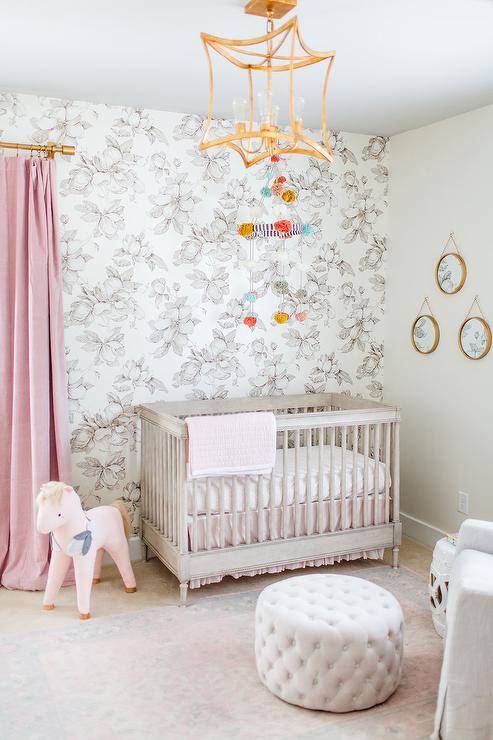 A brass lantern illuminates a stunning pink, white, and gray nursery. Pink curtains hang from a brass rod beside a gray French crib placed in front of a black and white floral wallpapered wall and beneath a pom pom mobile.