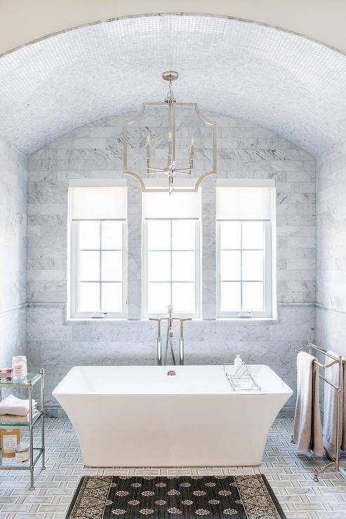 A Threshold Blue Block Print Braided Accent Rug sits on gray maze floor tiles in front of a Free Standing Cast Iron Soaking Bath Tub. A Wide Chandelier hung from a marble tiled barrel ceiling over the tub and in front of windows covered in white roman shades. The bathtub is flanked by a nickel and glass tiered accent table and a Towel Stand.