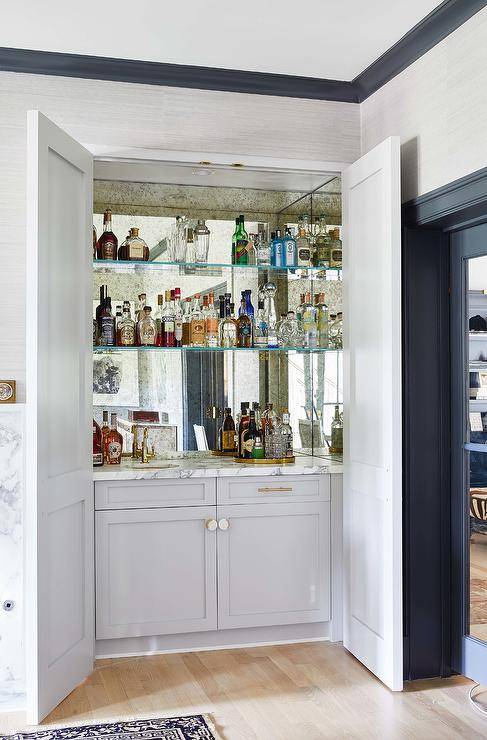 Hidden behind light gray double doors, glass shelves are mounted against a smoked mirror backsplash and over light gray shaker cabinets finished with a round sink and a polished brass faucet.