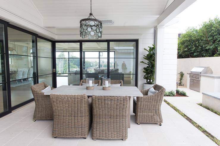 A recycled glass discs chandelier hangs from a white wood plank patio ceiling over a concrete dining table surrounded by brown wicker dining chairs.