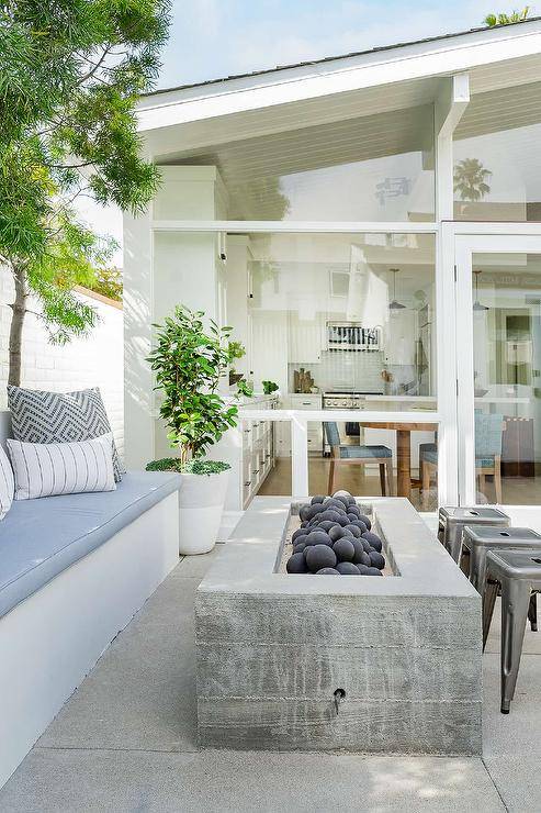 Patio features an outdoor built in sofa adorned with blue and gray cushions and a rectangular concrete fire pit with tolix stools.