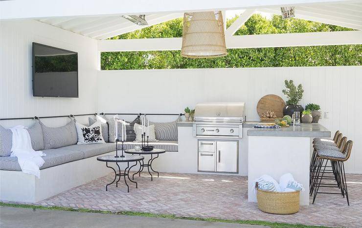 Transitional outdoor patio featuring a white built-in outdoor sofa with gray upholstered cushion and rattan counter stools. Red brick herringbone pattern pavers finish the surface with a timeless rustic appeal.