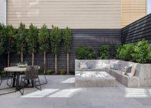 A built-in l-shaped concrete sofa sits in a backyard at a concrete firepit, while gray rope chairs surround a concrete and metal dining table positioned on concrete pavers.