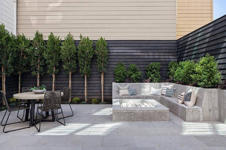 A built-in l-shaped concrete sofa sits in a backyard at a concrete firepit, while gray rope chairs surround a concrete and metal dining table positioned on concrete pavers.