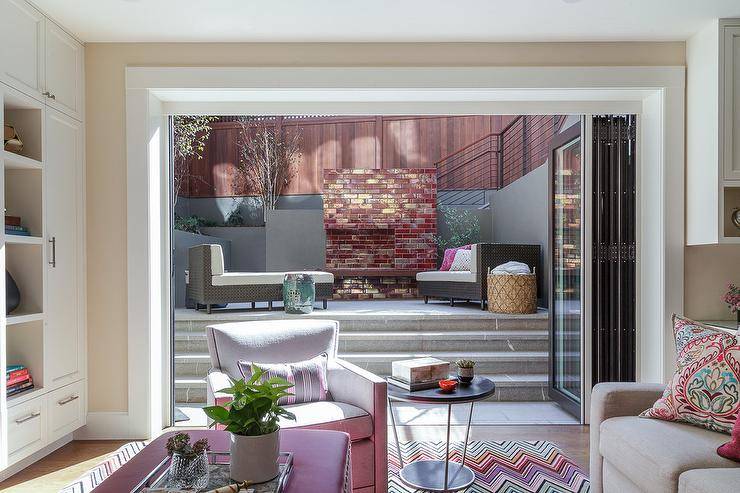 Folding glass doors open to a patio fitted with concrete patio steps leading to wicker loungers placed facing a red brick fireplace.
