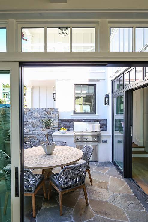 An outdoor kitchen fitted with a light gray concrete countertop is positioned to the side of sliding glass doors and in front of a round wood dining table seating Candelabra Home Loom Dining Chairs.