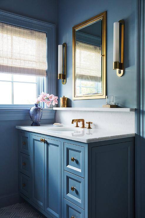 Brass and glass cylinder sconces mounted to a blue wall flank a brass framed inset medicine cabinet fixed above a blue washstand. The washstand dons aged brass knobs and an aged brass faucet fixed to a marble countertop under a marble shelf mounted against a marble backsplash.