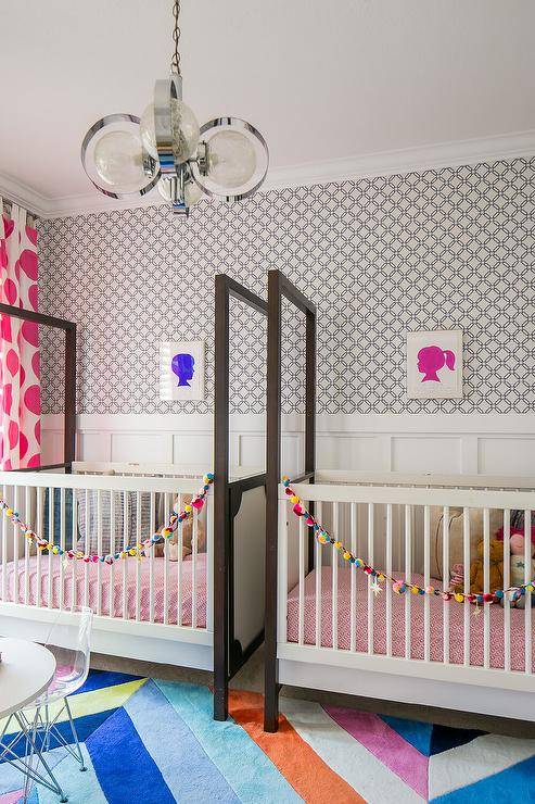 Contemporary nursery for fraternal twins boasts side-by-side white and brown cribs placed on a colorful geometric rug underneath silhouette art pieces hung from a wall clad in undecorous geometric wallpaper lined white workbench and spoke trim.