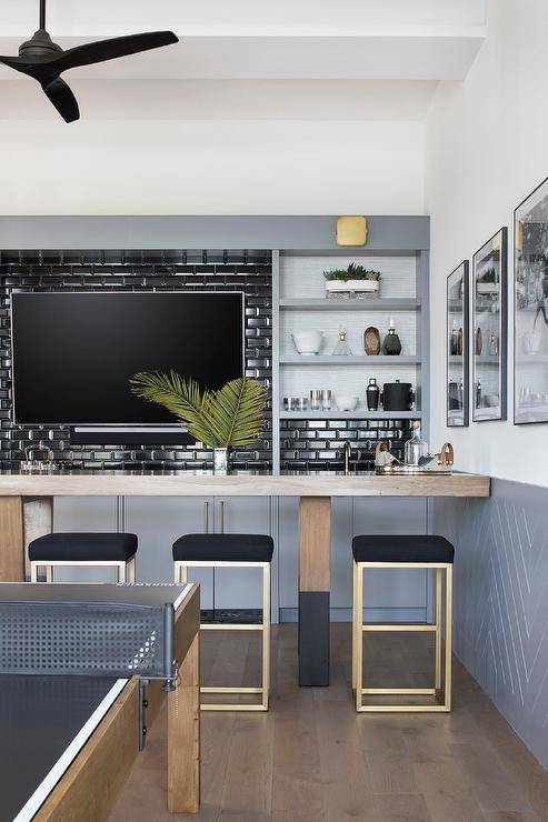 Contemporary wet bar features a TV mounted on black beveled subway tiles, a brown and black bar with gold and black barstools and built in shelves.