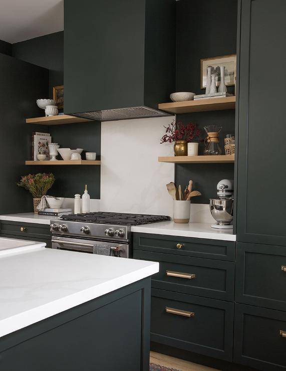 Black and white kitchen features blond floating shelves, a black hood over a white marble slab backsplash and black cabinets with brass pulls.