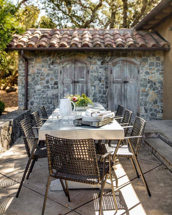 Outdoor patio designed with a concrete dining table and black woven chairs atop large concrete pavers. This rustic outdoor space features cobblestone surroundings and distressed wood doors for a charming outdoor escape.