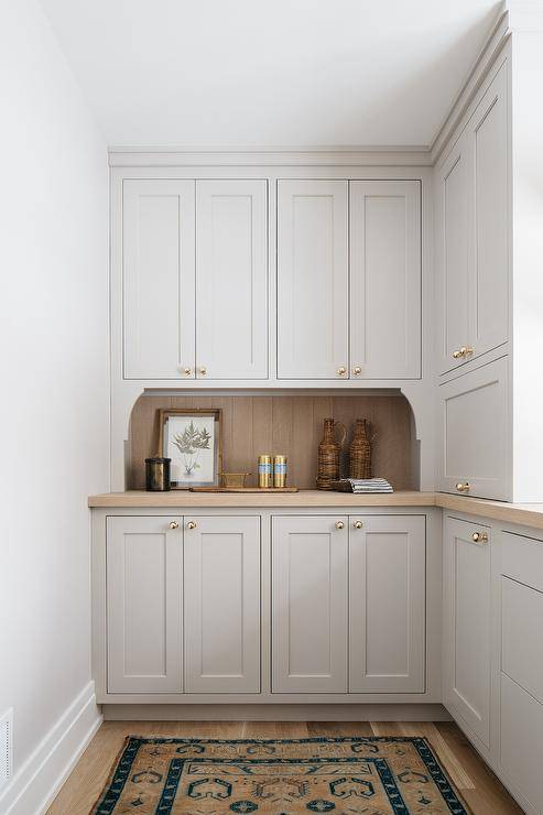 A wood plank backsplash accents light gray pantry cabinetry donning vintage brass ball knobs and a wooden countertop. A beige and blue vintage rug sits in front of the cabinets on a wood floor.