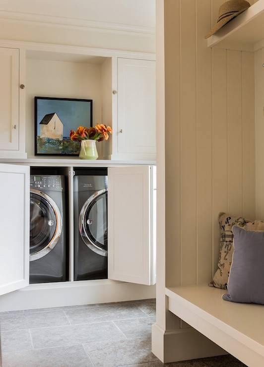 Country laundry room features a concealed washer and dryer hidden behind folding doors under a shelf filled with art and a two tone pitcher filled with flowers situated across from a built-in bench lined with vertical paneling.
