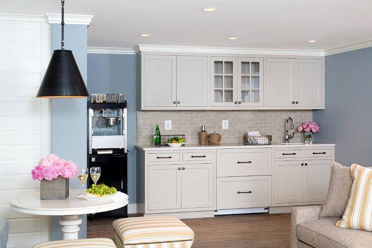 Beautifully designed basement kitchenette features gray shaker cabinets accented with oil rubbed bronze pulls and an ivory quartz countertop fitted with a round sink and polished nickel faucet fixed in front of gray stacked backsplash tiles. A glass front upper cabinet is flanked by gray cabinets beautifully complementing blue walls. The kitchenette is completed with a popcorn machine.