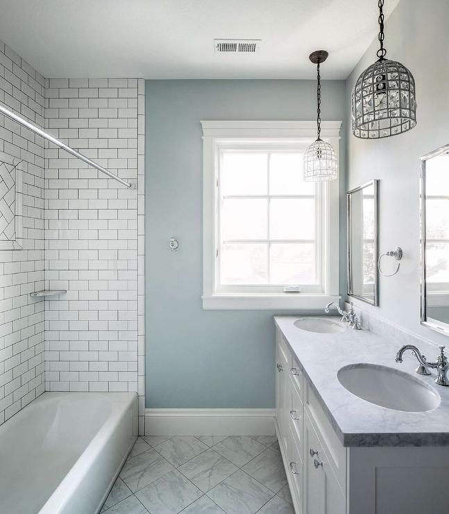 A pair of vintage glass birdcage chandelier hang over a double bath vanity topped with honed gray marble fitted with his and hers sinks placed under metal framed mirrors. A marble diamond pattern floor leads to a drop-in tub clad in white subway tiles finished with dark grout.